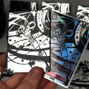 Può includere: Carta collezionabile nera e argento con un'illustrazione di guerriero. La carta ha un effetto olografico e include testo giapponese e il numero 12. La carta fa parte di un gioco di carte collezionabili.