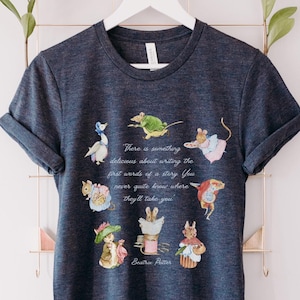 Puede incluir: Camiseta azul oscuro con ilustraciones de personajes de cuentos de hadas y la cita "Hay algo delicioso en escribir las primeras palabras de una historia. Nunca sabes a dónde te llevarán" de Beatrix Potter.