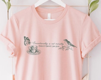 Könnte beinhalten: Ein hellrosa T-Shirt mit Rundhalsausschnitt und kurzen Ärmeln. Das Shirt zeigt eine Grafik eines Schmetterlings, einer Teetasse und eines Vogels mit dem Text "Conscientiously is not morality. Others haven't just fine."