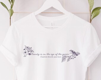 Könnte beinhalten: Weißes T-Shirt mit floralem Design und dem Zitat "Beauty is in the eye of the gazer" von Charlotte Brontë, Jane Eyre. Das Design ist in Lila- und Grautönen gehalten. Das T-Shirt hängt an einem weißen Kleiderbügel.