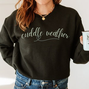May include: A black sweatshirt with the text "cuddle weather" in a light gray font with a small heart symbol.
