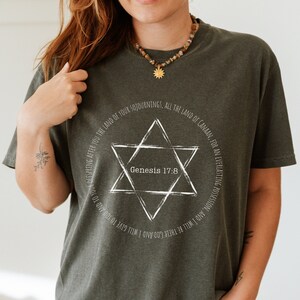 Könnte beinhalten: Dunkel olivgrünes T-Shirt mit einem weißen Davidstern-Design und dem Text "Genesis 17:8". Das Design ist von Schrift umgeben. Das Shirt ist aus weichem Material.