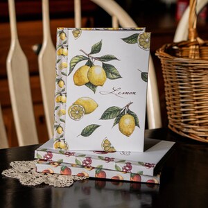 May include: A stack of three notebooks with a lemon theme. The top notebook has a white cover with a lemon branch design and the word "Lemon" written in cursive. The bottom two notebooks have a red and white floral design.