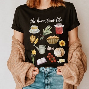May include: Black t-shirt with the text "the homestead life" printed in white. The shirt features a variety of illustrations of items associated with homesteading, including a loaf of bread, a jar of jam, a watering can, a book, and a bunch of green onions.