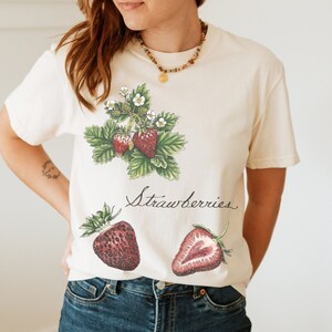 Puede incluir: Una camiseta blanca con un gráfico de fresas y la palabra "Strawberries" impresa en ella. La camiseta la lleva puesta una persona con el pelo largo y castaño.