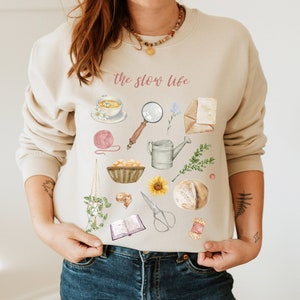 May include: A beige sweatshirt with the text "the slow life" printed in pink. The sweatshirt features a variety of illustrations, including a cup of tea, a magnifying glass, a watering can, a book, yarn, a sunflower, and a loaf of bread.