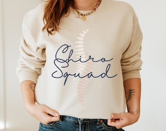 Pode incluir: Uma camisa de moletom bege com um gráfico de coluna vertebral e o texto "Chiro Squad" em escrita azul marinho.