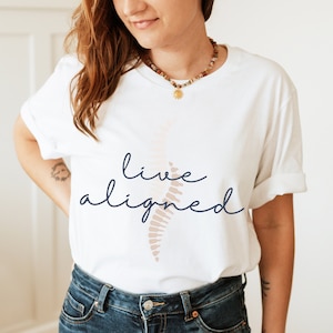 May include: White t-shirt with the words "live aligned" in navy blue script and a beige spine graphic. The shirt is made of a soft material and has rolled-up sleeves. The overall theme is wellness and self-care.