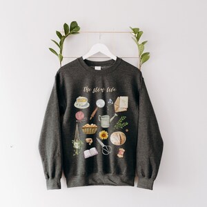 May include: A dark gray sweatshirt with the text "The Slow Life" printed on the front. The design features a variety of illustrations, including a cup of tea, a book, a watering can, a loaf of bread, and a sunflower.