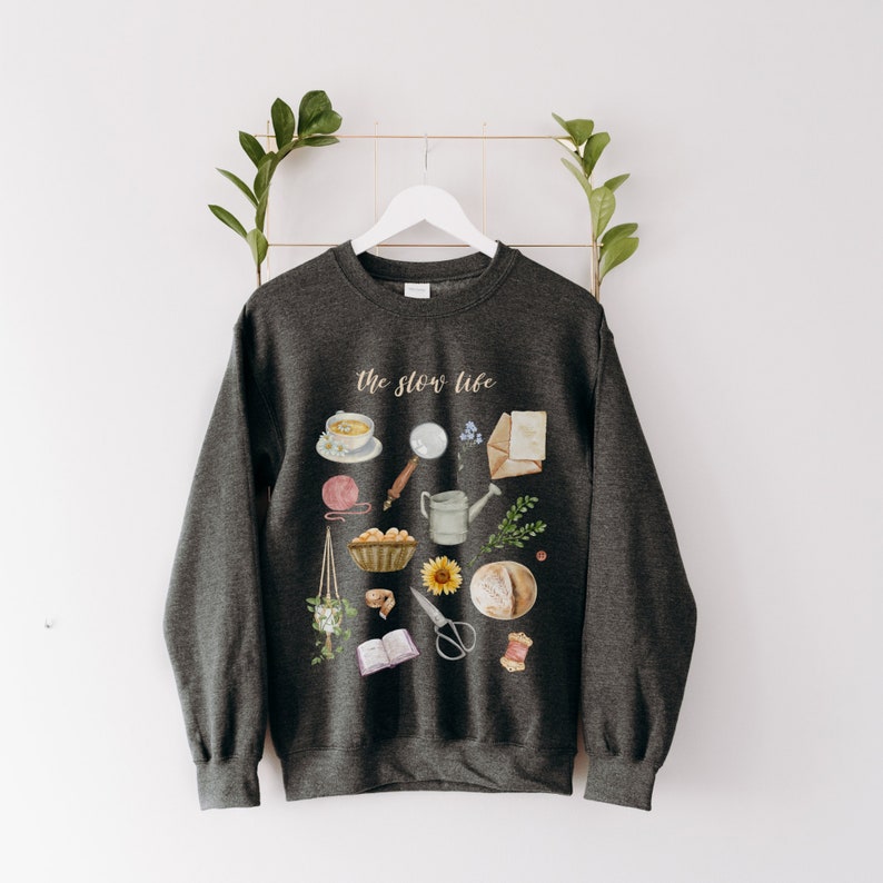 May include: A dark gray sweatshirt with the text "The Slow Life" printed on the front. The design features a variety of illustrations, including a cup of tea, a book, a watering can, a loaf of bread, and a sunflower.