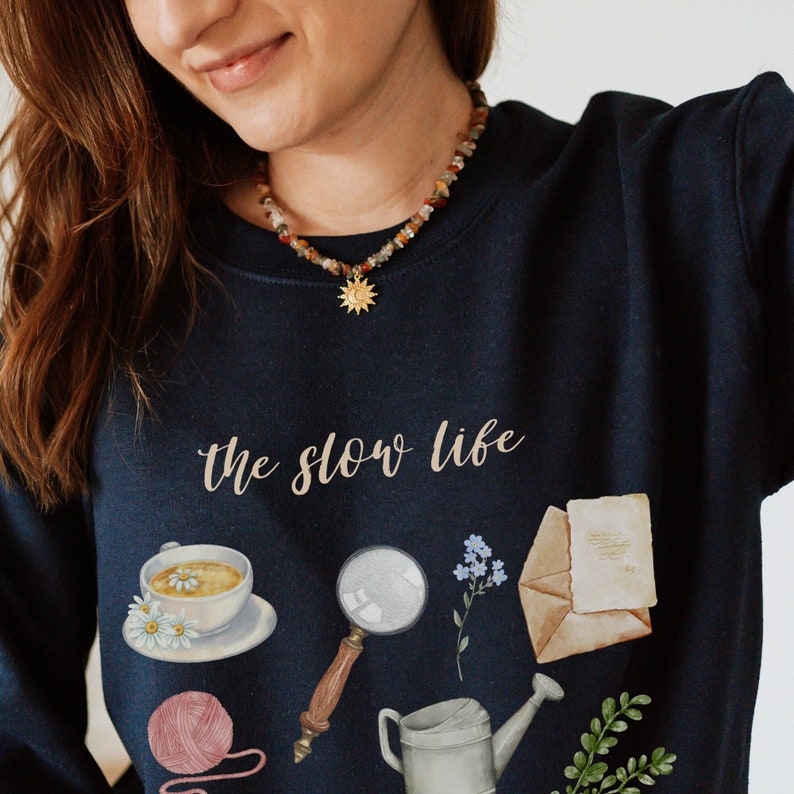 May include: A navy blue sweatshirt with the text "the slow life" printed on the front. The sweatshirt features a variety of illustrations, including a cup of tea, a magnifying glass, a watering can, a ball of yarn, and a bouquet of flowers.
