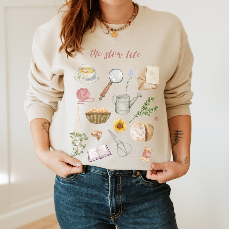 May include: A beige sweatshirt with the text "the slow life" printed in pink. The sweatshirt features a variety of illustrations, including a cup of tea, a magnifying glass, a watering can, a book, yarn, a sunflower, and a loaf of bread.