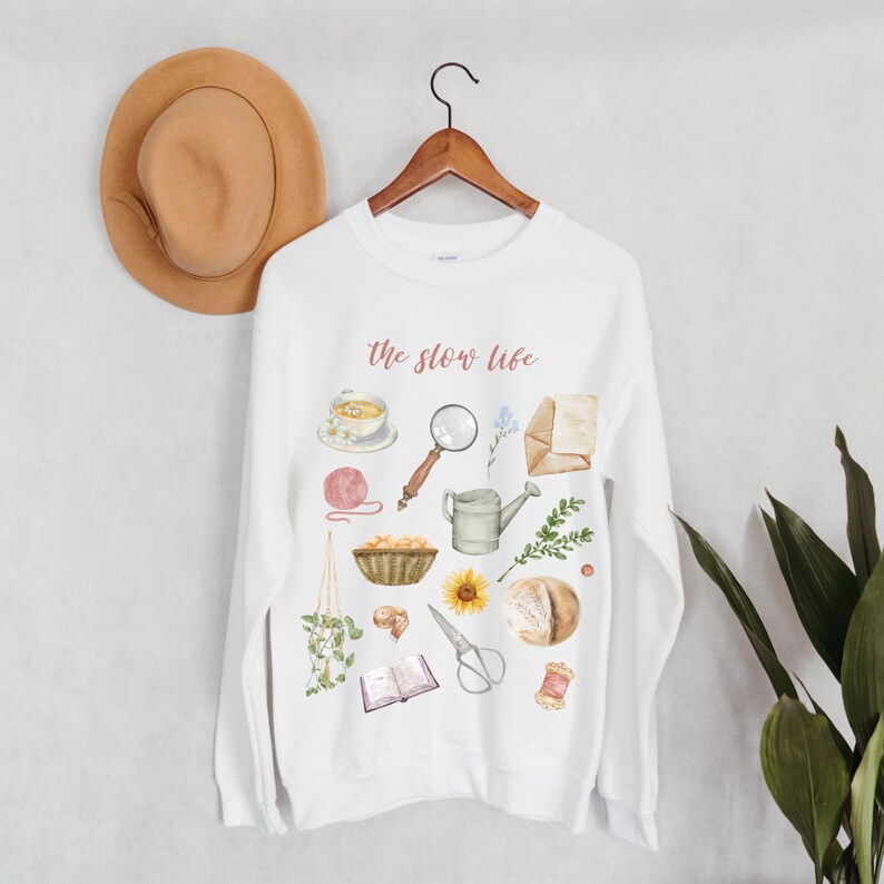 May include: White sweatshirt with the text "the slow life" printed in pink. The sweatshirt features a variety of watercolor illustrations of items associated with a slow lifestyle, including a cup of tea, a magnifying glass, a watering can, a loaf of bread, a book, and a sunflower.