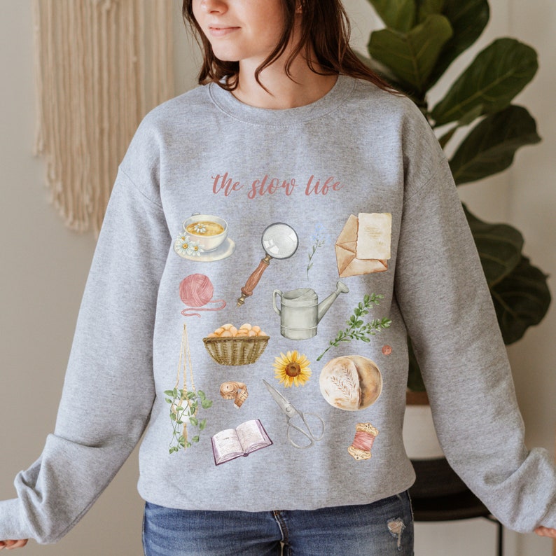 May include: Gray sweatshirt with the text "the slow life" printed in pink. The sweatshirt features a variety of illustrations, including a cup of tea, a magnifying glass, a watering can, a sunflower, a loaf of bread, a book, and a spool of thread.