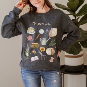 May include: A dark gray sweatshirt with the text "the slow life" printed on the front. The sweatshirt features a watercolor illustration of various items, including a cup of tea, a magnifying glass, a watering can, a loaf of bread, a sunflower, and a book.