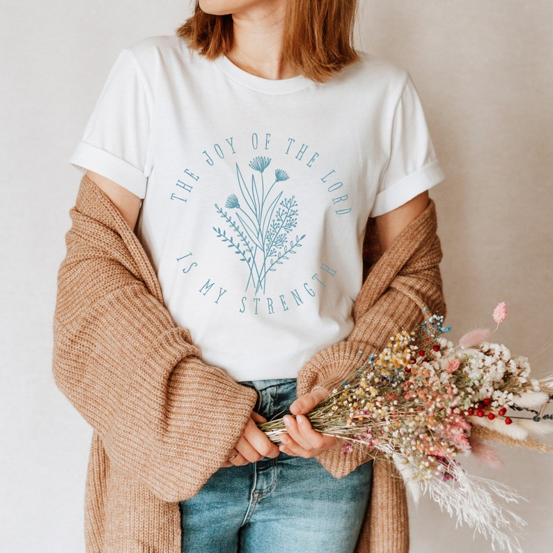 May include: White t-shirt with a blue floral design and the text "The Joy of the Lord is My Strength".