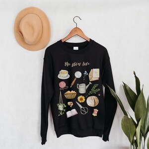 May include: Black sweatshirt with the text "the slow life" in white lettering. The sweatshirt features a variety of illustrations in watercolor style, including a cup of tea, a magnifying glass, a watering can, a sunflower, a loaf of bread, and a book.