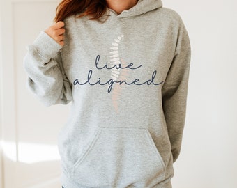 Op de afbeelding: Grijs hoodie met de tekst "live aligned" in donkerblauwe schrift. Achter de tekst bevindt zich een gestileerd roze en wit varenbladontwerp.