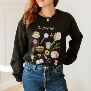 May include: Black sweatshirt with a white graphic print of various items, including a cup of tea, a magnifying glass, a watering can, a sunflower, a loaf of bread, and a book. The text "the slow life" is printed in white on the sweatshirt.