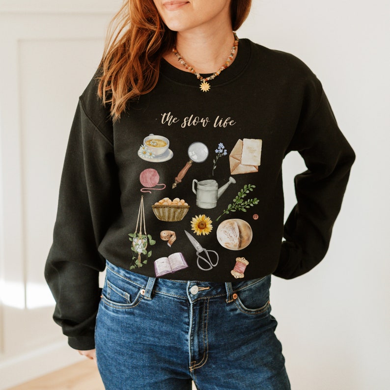 May include: Black sweatshirt with a white graphic print of various items, including a cup of tea, a magnifying glass, a watering can, a sunflower, a loaf of bread, and a book. The text "the slow life" is printed in white on the sweatshirt.