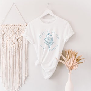 May include: White t-shirt with a blue floral design and the text "The Joy of the Lord is My Strength".