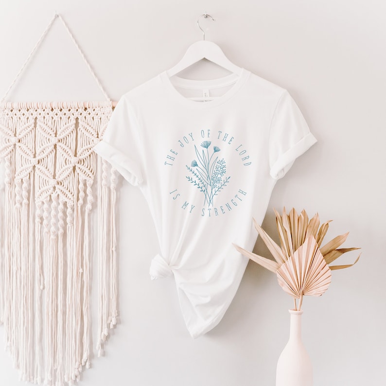 May include: White t-shirt with a blue floral design and the text "The Joy of the Lord is My Strength".