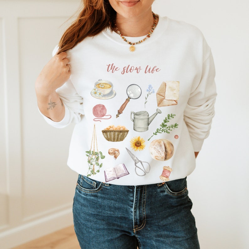 May include: White sweatshirt with the text "the slow life" printed on the front. The sweatshirt features a variety of illustrations, including a cup of tea, a magnifying glass, a watering can, a basket of bread, a book, and a pair of scissors.