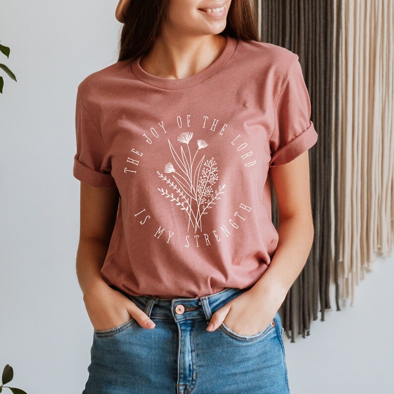 May include: A mauve t-shirt with a white graphic design featuring a floral arrangement and the text "The Joy of the Lord is My Strength".