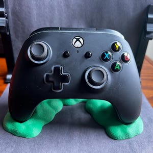 Puede incluir: Un mando Xbox negro con soportes verdes. El mando tiene una cruceta negra y blanca, botones negros y blancos y gatillos negros y blancos. El mando está apoyado en un soporte verde.