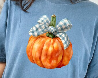 May include: A blue t-shirt featuring a watercolor-style pumpkin design. The pumpkin is orange with a green stem and a blue and white checkered bow. The shirt is a solid blue color.
