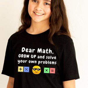 May include: Two girls wearing black t-shirts with the text "Dear Math, GROW UP and solve your own problems" and a smiley face with sunglasses. The text is surrounded by colorful math symbols.