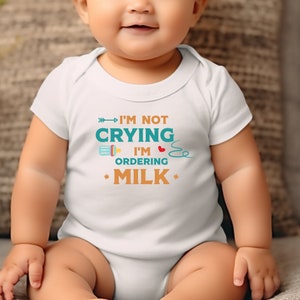 May include: White baby grow with a turquoise and orange graphic that says "I'm not crying I'm ordering milk" with a baby bottle and a heart.