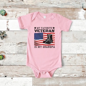 May include: Pink baby grow with a graphic of an American flag and boots. The text reads "My Favourite Veteran is My Grandpa."