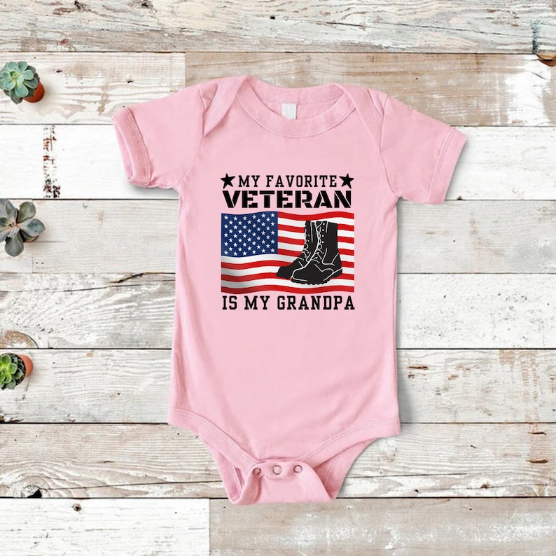 May include: Pink baby grow with a graphic of an American flag and boots. The text reads "My Favourite Veteran is My Grandpa."