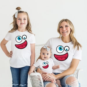 May include: A family of three wearing white t-shirts with a cartoon face design. The design features a large blue eye, a smaller blue eye, and a red smiling mouth. The family is posing for a photo, with the mother holding the baby in her arms.