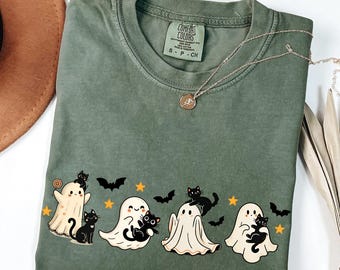May include: Olive green Comfort Colors t-shirt featuring a Halloween design. The design includes three cartoon ghosts, black cats, bats, and stars. A gold necklace with a pendant is also visible.