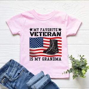 May include: Pink t-shirt with a black and white American flag, black boots, and the text "My Favourite Veteran is My Grandma".