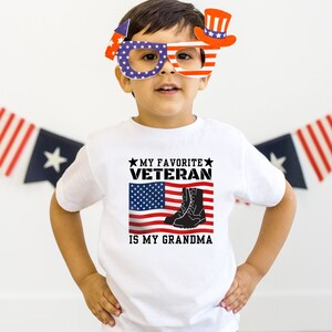 May include: A white t-shirt featuring a red, white and blue American flag design. The text "My Favorite Veteran Is My Grandma" is printed on the shirt. The shirt is being worn by a young child wearing a red, white and blue paper hat and glasses.