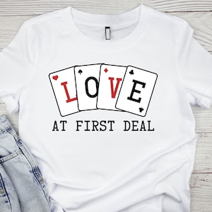 May include: White t-shirt with a graphic of playing cards spelling out "LOVE" and the text "AT FIRST DEAL".