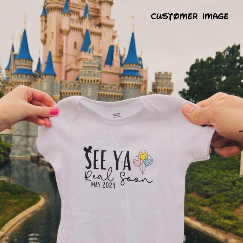 May include: White baby onesie with black text that reads "See Ya Real Soon May 2024" and a Mickey Mouse head with three colorful balloons. The onesie is held in front of a pink castle with blue towers.