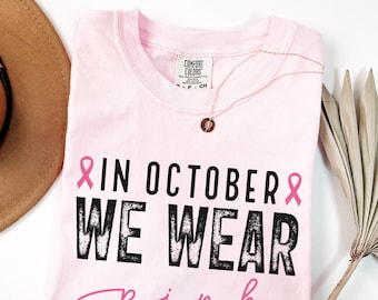 May include: A light pink t-shirt with the text "IN OCTOBER WE WEAR pink" in black and pink. A gold necklace with a pendant is draped on the shirt. A brown hat and dried palm leaf are in the background.