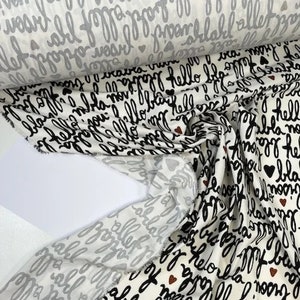 May include: White fabric with a repeating pattern of black cursive text, including the word "hello" and other phrases. Small black and brown hearts are scattered throughout the design. A roll of the fabric is visible in the background.