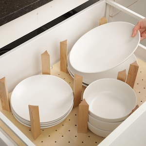 May include: A white kitchen drawer with wooden dividers holding stacks of white plates and bowls. A hand is placing a plate into the drawer. The drawer is inside a white cabinet with a black countertop.