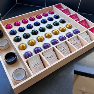 Puede incluir: Un organizador de madera con compartimentos para cápsulas de café y bolsitas de té. El organizador tiene una cuadrícula de 36 compartimentos para cápsulas de café y una fila de 6 compartimentos para bolsitas de té. Las cápsulas de café son de varios colores, incluyendo rojo, azul, verde y dorado. Las bolsitas de té están en paquetes blancos y rosados. El organizador está hecho de madera clara y tiene un acabado natural.