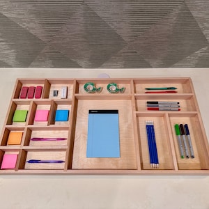 Puede incluir: Una bandeja organizadora de madera con múltiples compartimentos para guardar artículos de oficina. La bandeja contiene una variedad de artículos, incluyendo notas adhesivas, bolígrafos, lápices, borradores y cinta adhesiva.