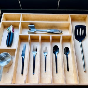 May include: A light-colored wooden drawer organizer with multiple compartments containing a silver meat tenderizer, a black slotted spatula, a stainless steel measuring cup, a garlic press, and a set of black cutlery including a knife, two forks, and two spoons.