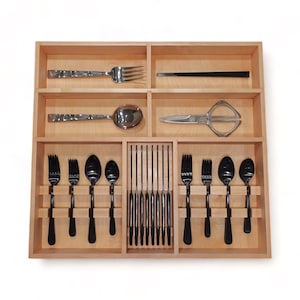 May include: A light-coloured wooden cutlery organiser tray with multiple compartments. It contains a silver fork, a pair of black chopsticks, a silver spoon, silver scissors, and a set of black forks and spoons arranged in organised slots.
