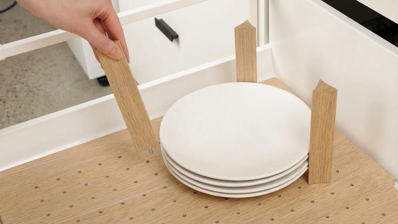 Może przedstawiać: Stos czterech białych ceramicznych talerzy w drewnianym stojaku na talerze. Stojak wykonany jest z jasnego drewna i ma za zadanie utrzymywać talerze w pozycji pionowej. Ręka umieszcza drewnianą przegrodę.
