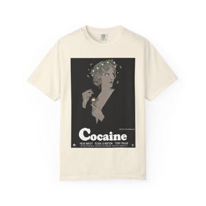May include: A cream-colored short-sleeve t-shirt featuring a black rectangular graphic print of a woman wearing a floral crown, holding a small object. The text reads Cocaine, A Master Team Presentation, Hilda Bayley, Flora Le Breton, Tony Fraser.