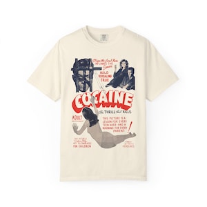 May include: A cream-colored short-sleeve t-shirt featuring a vintage-style movie poster graphic. The print includes the red text COCAINE, a central illustration of a person, and various black and red captions including The Thrill That Kills.